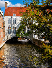Brugge, Belgium. Ancient medieval old town of Bruges. Sunny day. Channel arch bridge under house with red tegular roof and tree on the banks. Summer landscape with blue sky and clouds.