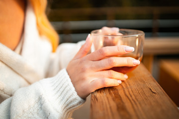 Close up of gentle woman hands with a glass cup of hot tea in the cold forest. Happy european blonde girl is keeping warm with hot tea in the cold autumn city park. Catch a cold...