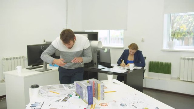 Man makes photo of model of houses, woman behind him sits and works with schemes