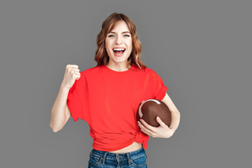 Freestyle. Woman in sports clothes studio standing isolated on grey with american football ball hand in fist up succeessful smiling happy