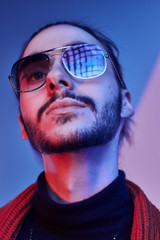 Bearded young man is posing for photographer under unusual light, wearing sunglasses.