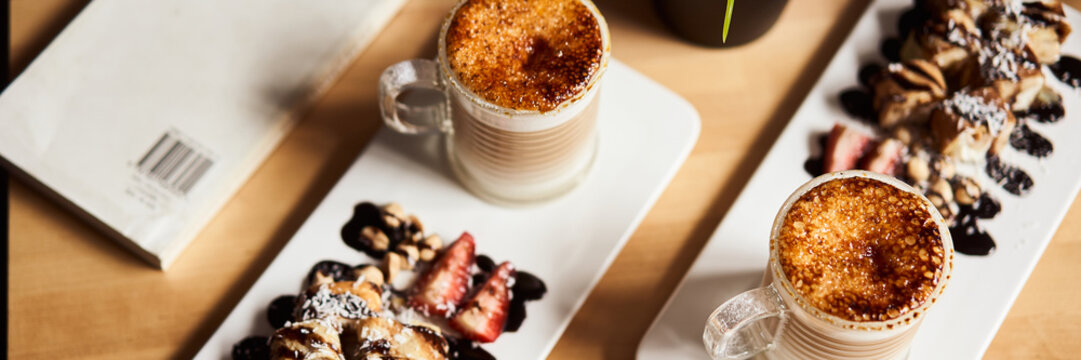 Banner size image of two cups of Hot latte with baked caramel crust and sweet roll with banana and strawberry on the wooden table in coffe-shop. Coffee concept