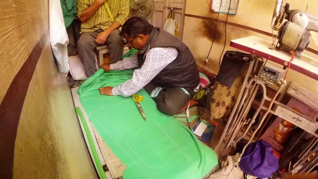 tailor draws pattern on green cloth on table old sewing-machine dirty walls Indian sewing workshop Bikaner