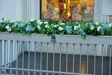 two plastic flowerpots on iron bars of a fence with decorative white and blue flowers on the street against the wall of the house with a window