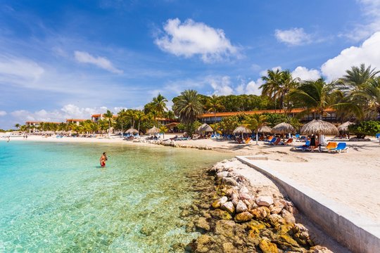 Beautiful view of white sand Curacao beach. Blue sunbeds under sun umbrellas on turquoise water and blue sky with white clouds background. Curacao Island.  Beautiful backgrounds.