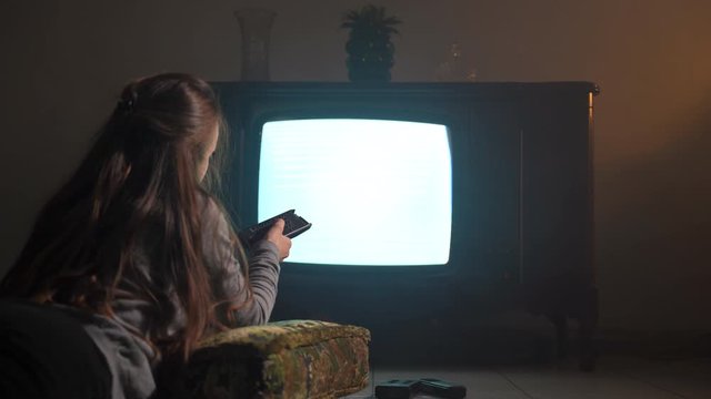 Girl lying on soft old-fashioned textile pillow on the floor in dark mist room and watching retro TV with flicking screen. Woman changing channels with TV remote control, horror atmospere and