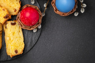 Painted Easter eggs in wicker coasters, pieces of Easter cake on a cutting board on a dark table - traditional Easter breakfast, top view, flat lay