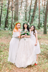 Fototapeta na wymiar Rustic wedding outdoors. The bride in a white dress and floral wreath standing with her two bridesmaids in green forest.