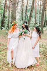 The group photo of pretty bride holding stylish rustic wedding bouquet and two bridesmaids in white dresses smiling and looking each other, standing in sunny green pine forest
