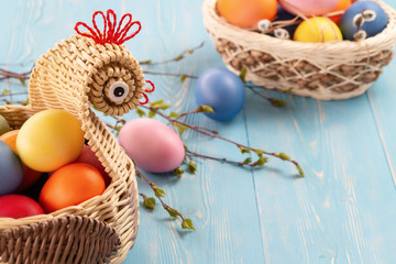 Wicker basket in the form of a chicken with colorful Easter eggs on blue wooden table - easter composition with copyspace