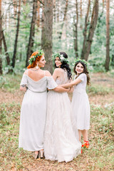 Back view of pretty cheerful bride and bridesmaids in white dresses and floal wreaths having fun at wedding day, walking in the pine forest.