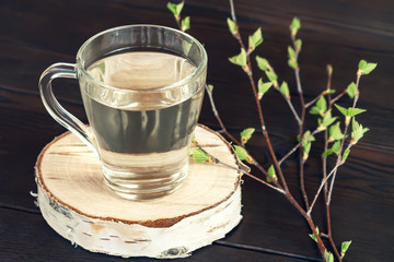 Birch juice on a dark table in a glass mug, next to a branch of birch with young leaves