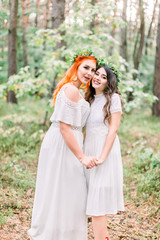 Two beautiful stylish happy bridesmaids in white dresses and wreaths on heads, smiling, holding hands and embracing, standing on the background of pine forest