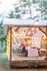 Blurred photo of wooden tent restaurant with wooden tables and chairs., decorated with light bulbs Vintage design of the restaurant in the forest in summer. Wedding reception in the open air.