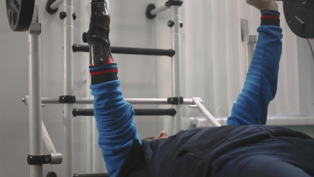 An athlete without a hand raises the barbell while lying down. Holds the bar with a professional sports hand prosthesis. Gym for Paralympics
