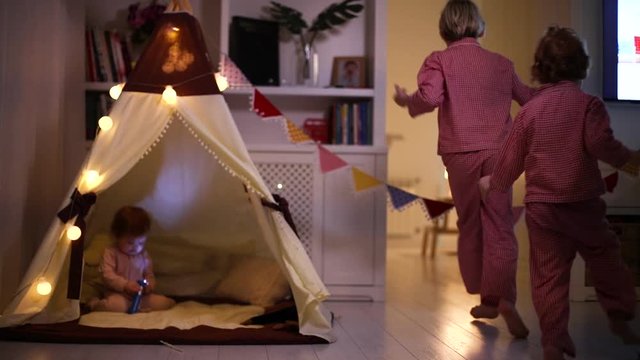 laughing kids, siblings having fun, running and playing around a teepee tent in the evening at home