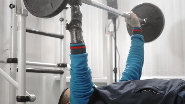 An athlete without a hand raises the barbell while lying down. Holds the bar with a professional sports hand prosthesis. Gym for Paralympics