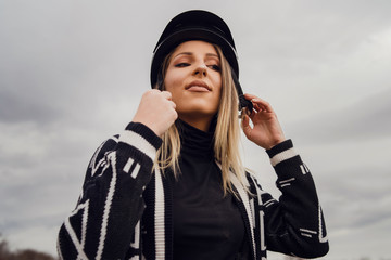 Young beautiful blonde caucasian woman female adjusting black protective helmet against a cloudy gray sky in winter or autumn day wearing sweater