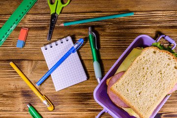 Different stationeries and lunch box with sandwiches on a wooden table. Top view