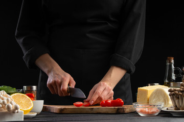 The chef cuts cherry tomatoes to prepare a tasty and fresh salad. Vegetarian and fresh food, gastronomy and cooking, recipe book and recipes. On a black background