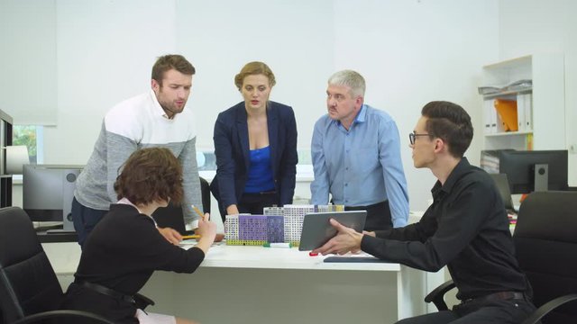 Young man sit at a table and show something on tablet to his workmates
