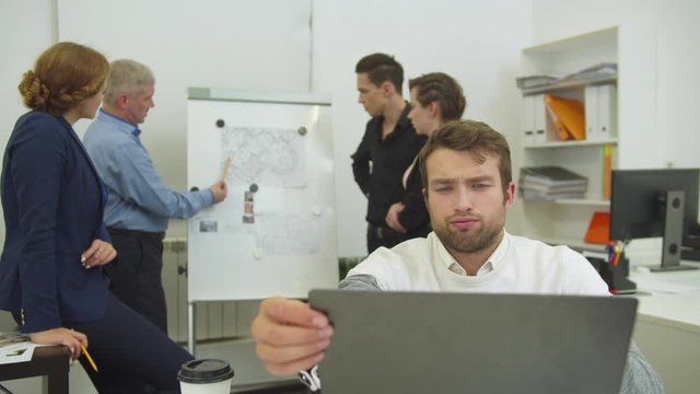 Tired man works at the laptop, his workmates discuss scheme of apartement on desk