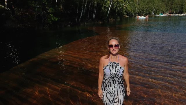 Woman walk on water on pier in sunglasses and a boho silk shawl. Girl rest on a flood wood underwater dock. The pavement is covered with water in the lake. In the background are mountain and a forest.