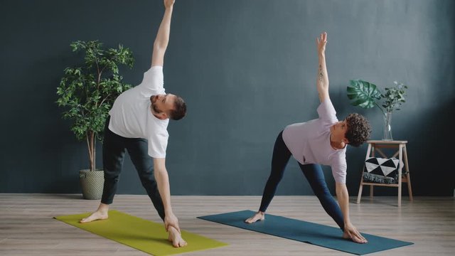 Male instructor bearded man is teaching female yoga student in cozy studio, young people are doing asanas enjoying healthy activity indoors. People and leisure concept.