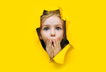 A little cute girl with red hair looks out through a hole in torn yellow paper and covers her mouth...