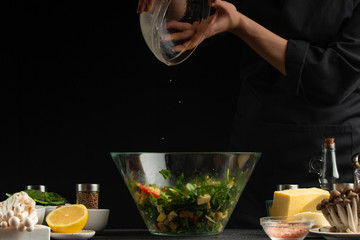 Cooking a salad, a cook preparing a salad on a dark background with ingredients, pouring sauce or dressing, freezing in motion. Delicious and healthy food. With space for design.