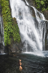 Young slim brunette woman with curly hair enjoying in lagoon of huge tropical waterfall Banyumala in Bali. Wearing in black swimsuit. Happy vacations in Indonesia. Wanderlust travel concept.