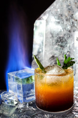 Yellow and orange drink with an ice sphere and green garnish with a flame next to it with a crystal in the background on an icey colored table top