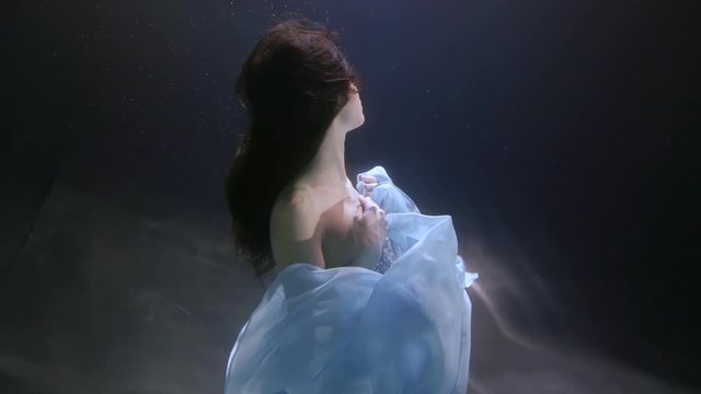 woman is sitting on bottom underwater and moving hands up, wearing silk dress