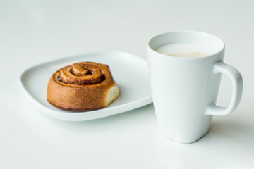 White cup of coffee with freshly baked cinnamon bun roll on a wihte plate. Minimalism