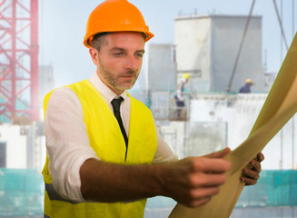 construction work lifestyle portrait of young happy and attractive engineer or building contractor holding blueprints wearing constructor hardhat smiling confident