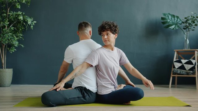 Slow motion of girl and guy enjoying pair yoga in wellness center sitting together on mat back to back doing asanas. Lifestyle and health care concept.