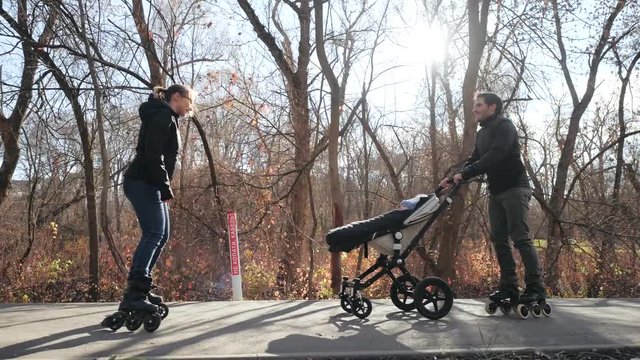 Sporty young family of skaters with a baby in a stroller in a cozy autumn park. Mom rides backwards and entertains the child. Slow motion.