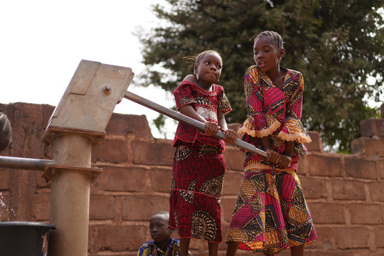 Young Girls Helping Each Other Pumping Water At An African Village Borehole