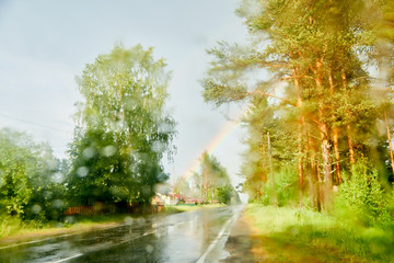 View from car cabin on wet asphalt road, trees and rainbow during rain and drops on front window