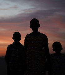 Silhouettes of Young African Girl With Her Younger Brothers Posing At A Pale Red Blue Sunset - obrazy, fototapety, plakaty