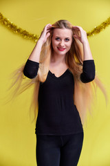 Naklejka premium Concept of a young blonde woman with New Year's decor. Portrait of a cute girl in a black T-shirt with long beautiful hair and great makeup. Smiling, showing emotions on a yellow background.