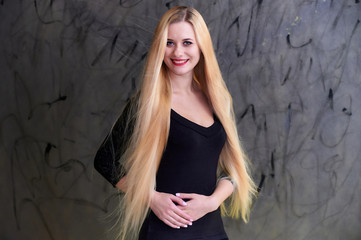 Fototapeta premium Concept of a young blonde woman with a chic hairstyle. Portrait of a cute girl in a black T-shirt with long beautiful hair and great makeup. Smiling, showing emotions on a gray background.