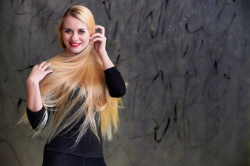Fototapeta premium Concept of a young blonde woman with a chic hairstyle. Portrait of a cute girl in a black T-shirt with long beautiful hair and great makeup. Smiling, showing emotions on a gray background.
