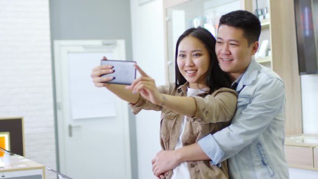 Handheld medium shot of cheerful young Asian couple taking selfie on new smartphone at gadget store, man hugging woman and smiling