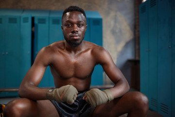 Fototapeta na wymiar Portrait of African young boxer looking at camera while sitting on bench in locker room