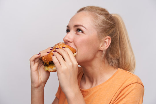 Close-up of young beautiful positive blonde woman with casual hairstyle delighting fresh tasty hanburger standing over white background in casual t-shirt