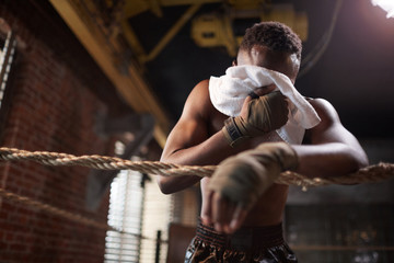Fototapeta na wymiar African muscular boxer standing on boxing ring and wiping face with towel after training in gym