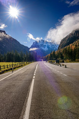 Fototapeta na wymiar Asphalt roads in the Italian Alps in South Tyrol, during autumn season / Sunny autumn day with dolomite mountains in background / Heavy sun flare thought the frame