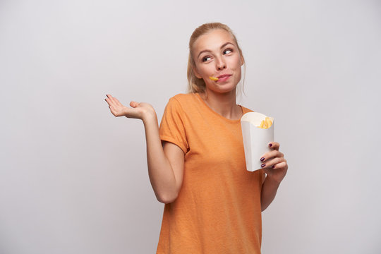 Indoor photo of positive young lovely blonde woman in casual clothes shrugging with raised palm and looking aside with cheerful smile, having french fries in her mouth
