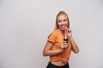 Studio photo of cheerful young attractive woman in orange t-shirt drinking soda with straw while looking aside with charming smile, posing against white background